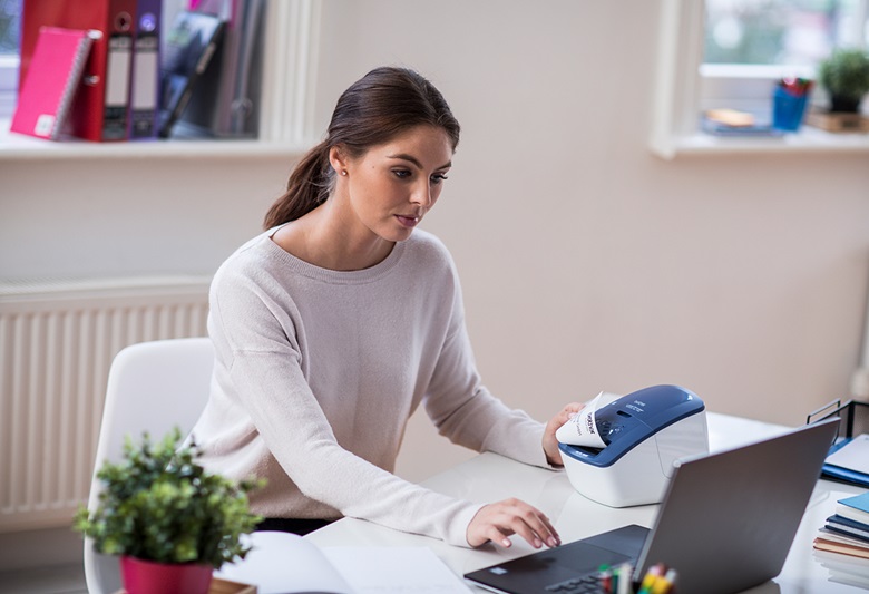 Žena sjedi i gleda u laptop koje ispisuje naljepnicu s uređaja s njezine lijeve strane.