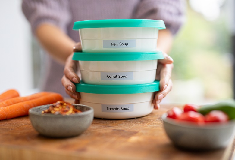 Žena drži tri označene kutije Tupperware na dasci za rezanje s mrkvom, rajčicama i krastavcima