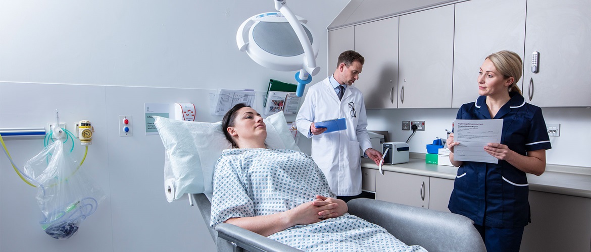 Pacijentica leži na krevetu, kraj nje medicinska sestra s obrascem, u pozadini liječnik u bijeloj uniformi